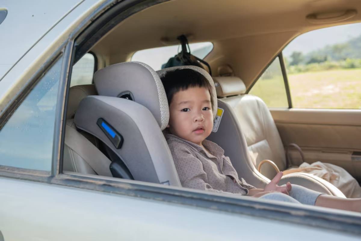 A child sits in a car's backseat, secured in a car seat, with scenic greenery visible through the open window.