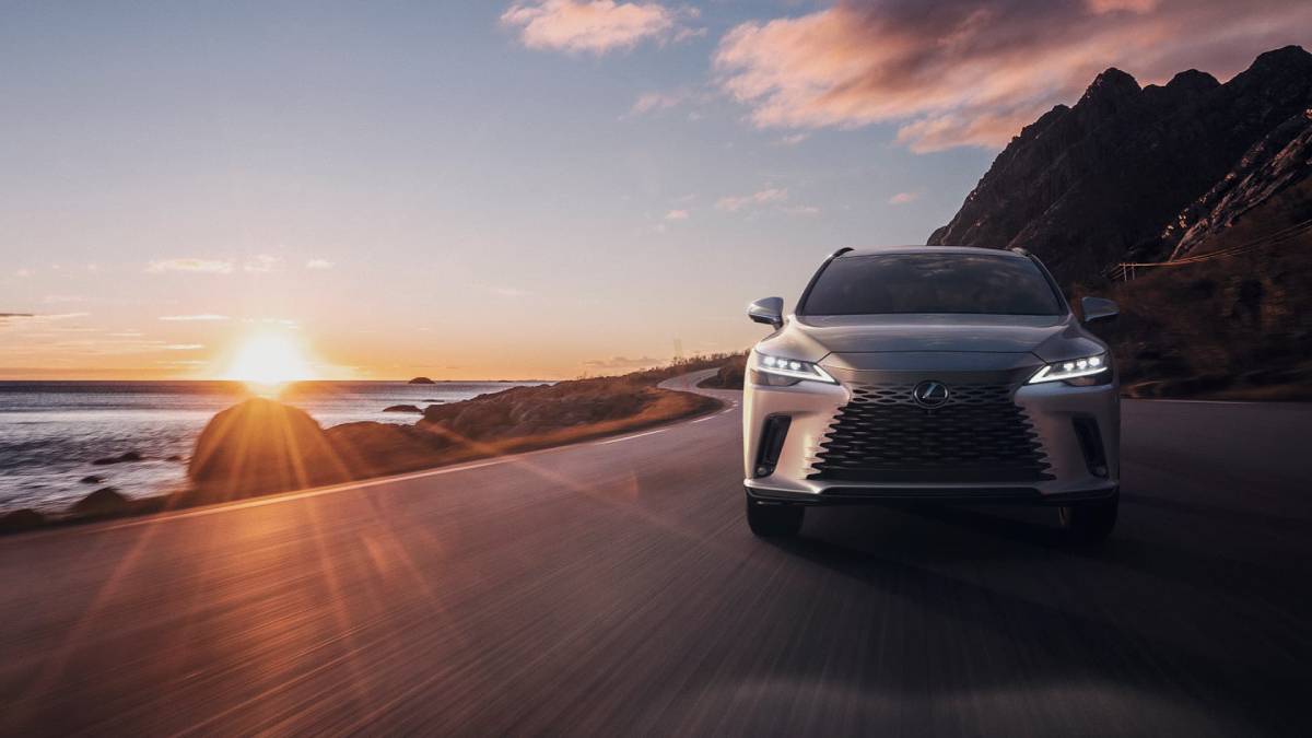 A silver Lexus SUV drives along a coastal road at sunset, with mountains in the background and the ocean glistening beside it.