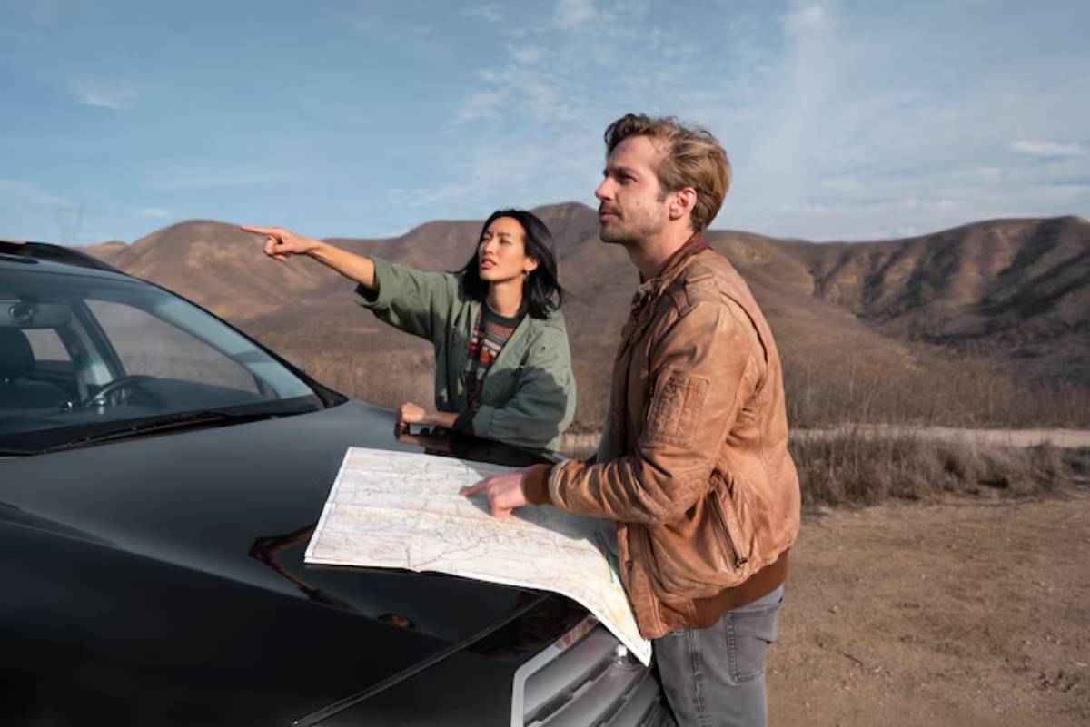 A man and woman examine a map on the hood of a black car, surrounded by mountains under a clear sky.