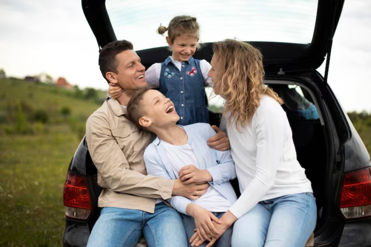 A happy family sitting together in the trunk of a car, enjoying a sunny day in a green, open landscape.