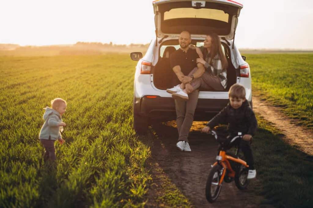 A family enjoys a sunny day outdoors, with kids playing near a parked car on a grassy field.