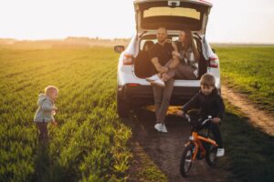 A family enjoys a sunny day outdoors, with kids playing near a parked car on a grassy field.