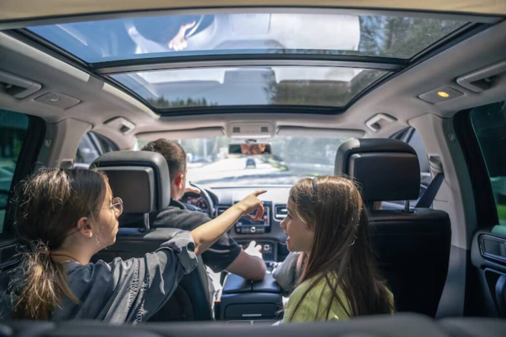 Inside a modern car, a person drives while two passengers engage in conversation, illuminated by sunlight streaming through the sunroof.