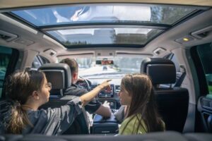 Inside a modern car, a person drives while two passengers engage in conversation, illuminated by sunlight streaming through the sunroof.