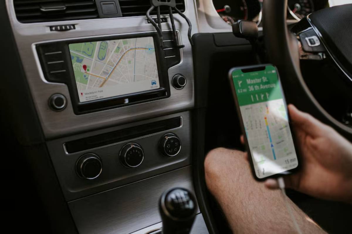 A driver's hand holds a smartphone displaying navigation directions, next to a car dashboard with a GPS map screen.