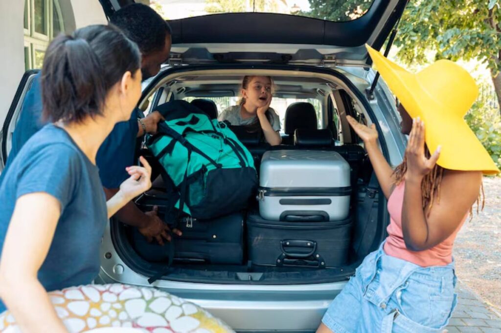 A group of friends pack luggage into the trunk of a car, preparing for a trip on a sunny day, with vibrant greenery in the background.