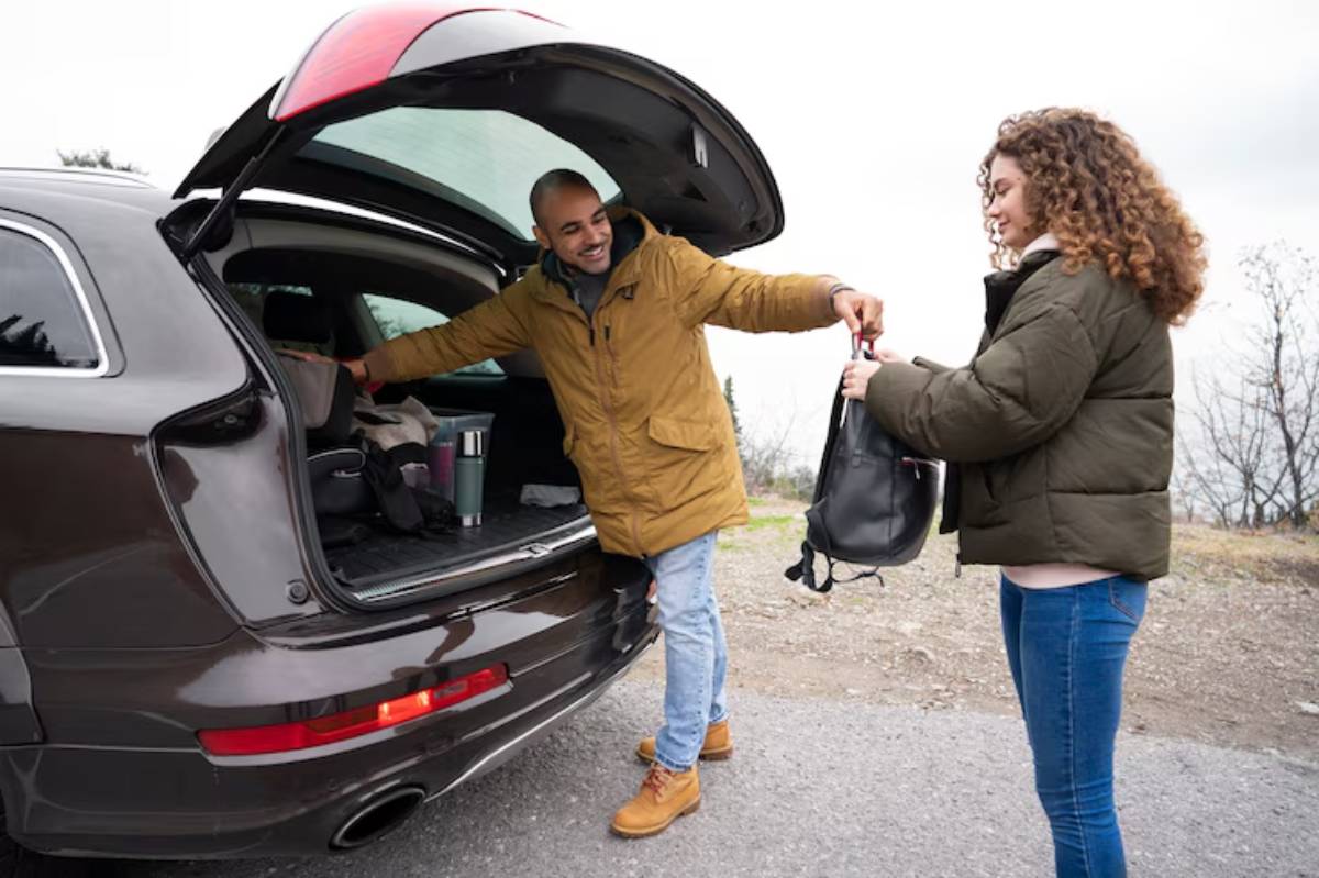 A man in a brown jacket hands a black bag to a woman in a green coat, near the open trunk of a dark SUV on a cloudy day.