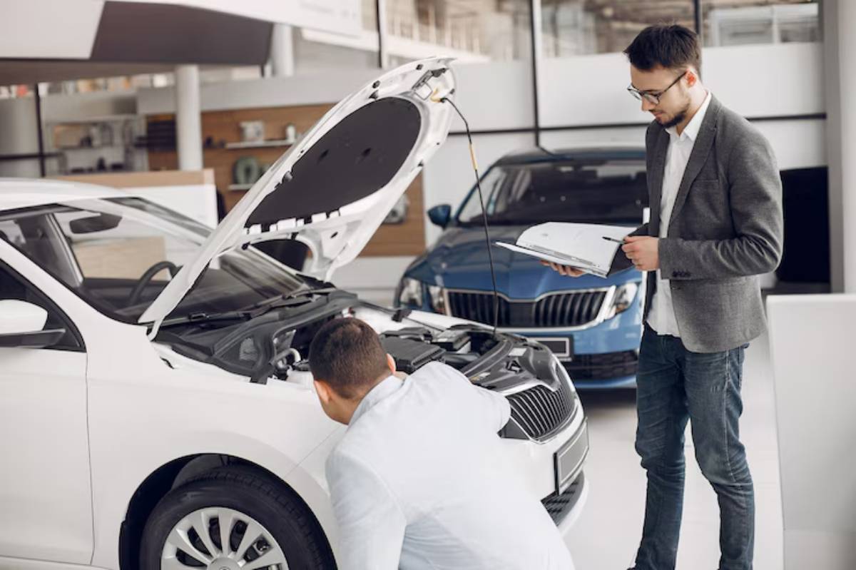A man inspects the engine of a white car in a dealership while another man holds a clipboard, observing the process.