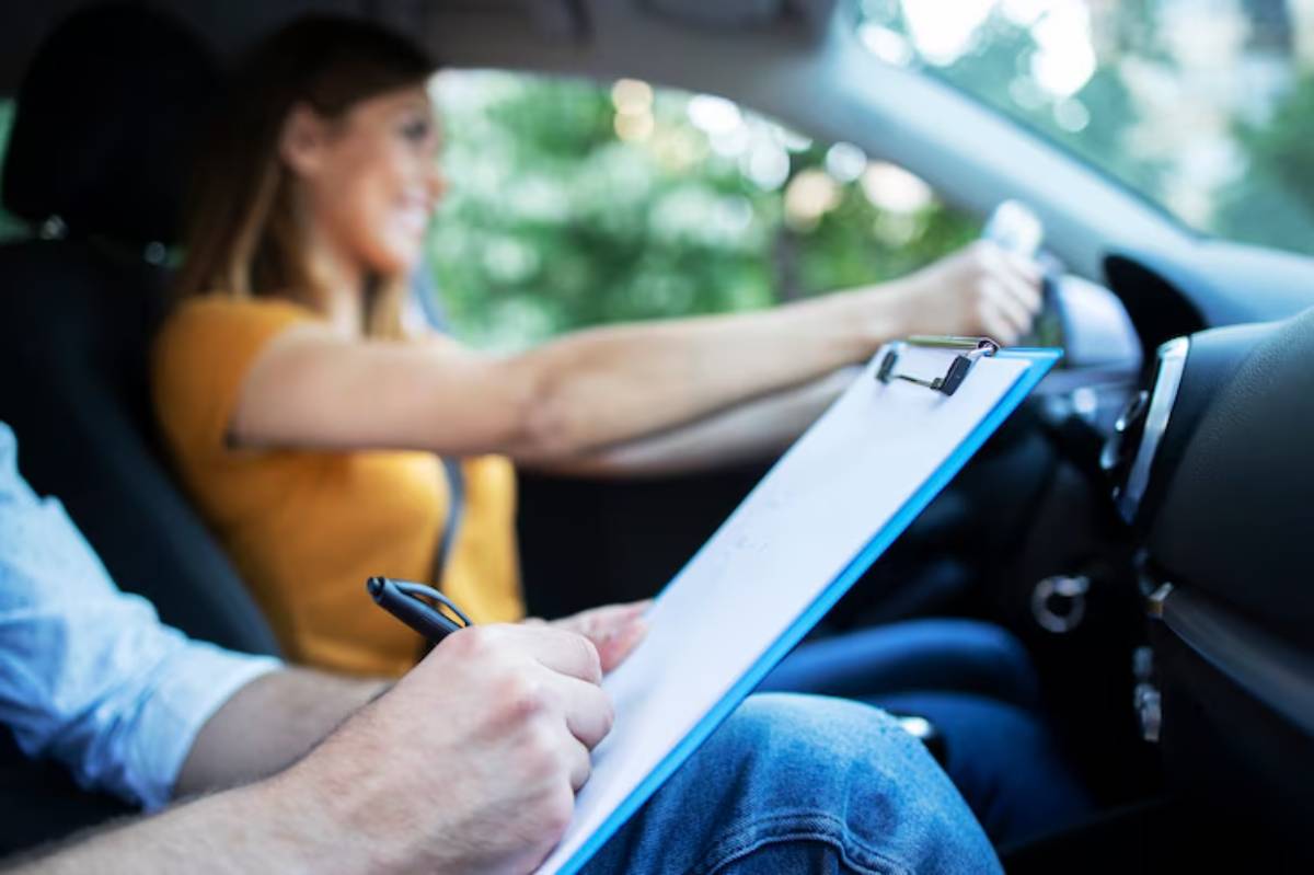 A seated person holding a clipboard and pen observes a learner driver at the wheel in a car during a driving lesson.