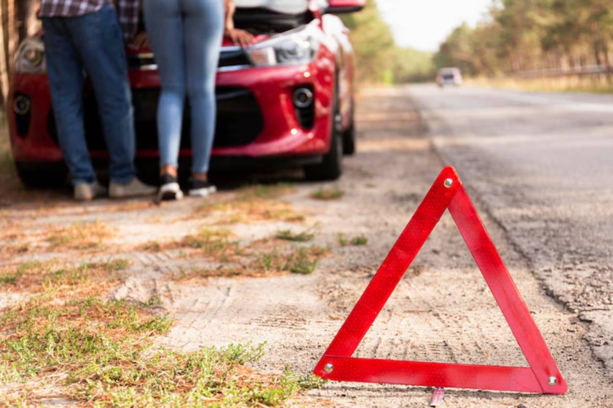 A red warning triangle on the roadside near a couple inspecting a car with its hood open, indicating a breakdown situation.
