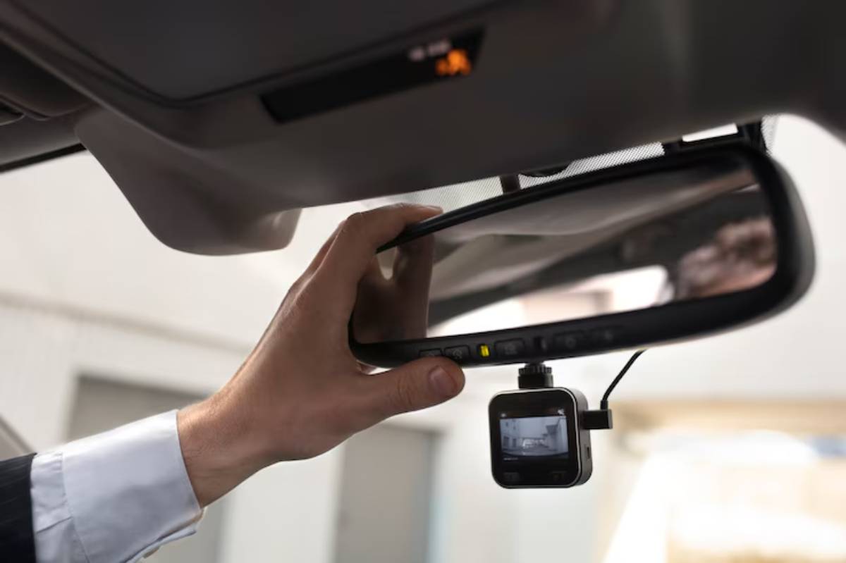 A hand adjusts a rearview mirror with an attached dash cam inside a vehicle. Clear camera display showing the view ahead.