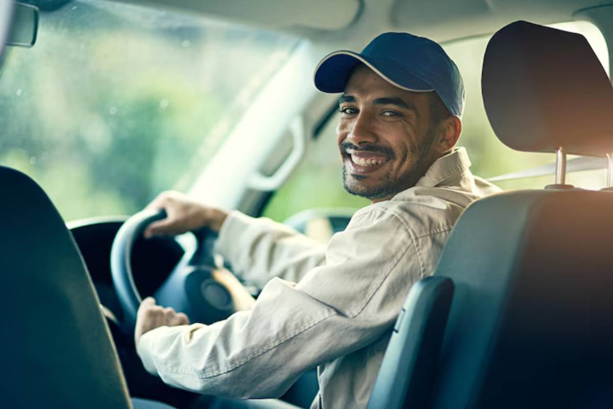 A man in a light jacket and cap drives a car, grasping the steering wheel, with a green landscape visible through the windshield.
