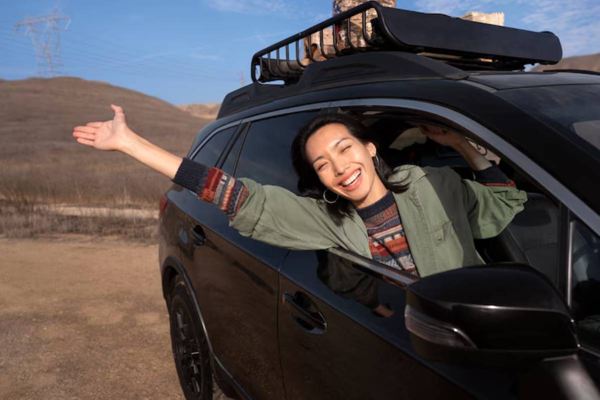 A person leans out of a black SUV parked in a rugged landscape, smiling and extending their arm towards the scenery.