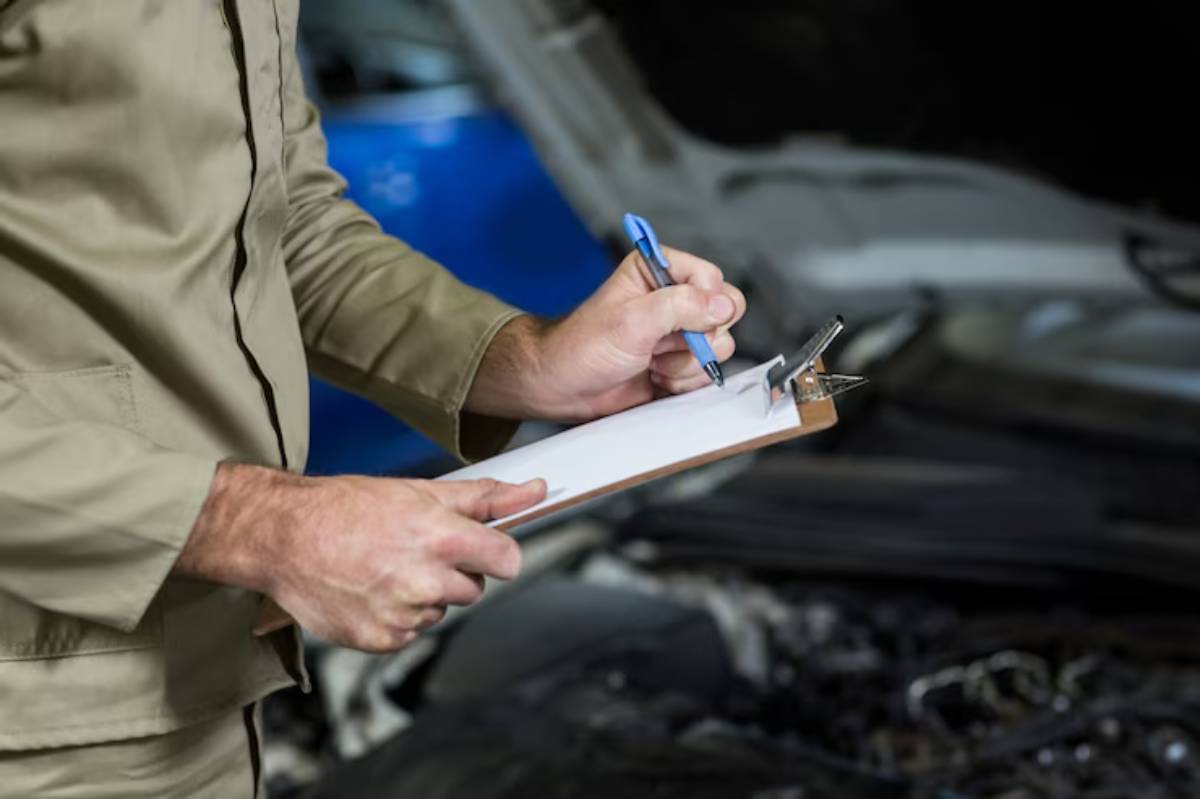 A mechanic in a beige uniform writes notes on a clipboard while inspecting a car's engine, focused on maintenance tasks.