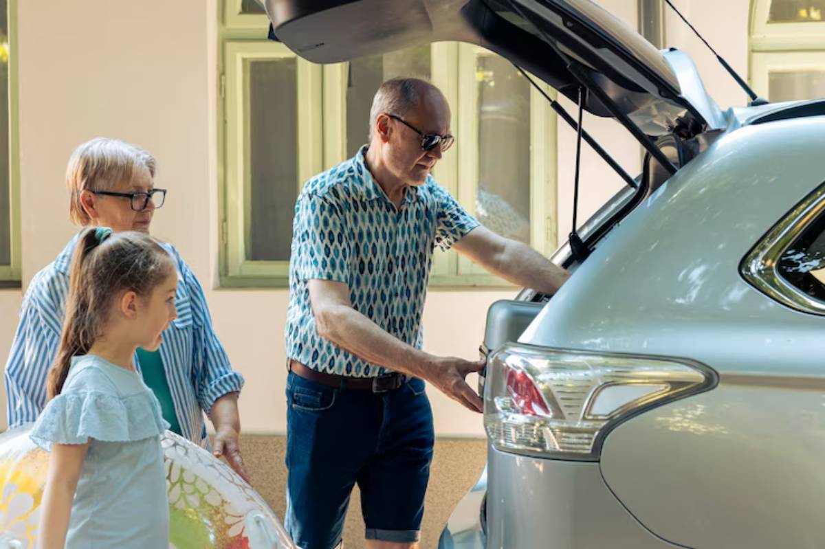 A man is loading a car trunk while a woman and a girl watch nearby, surrounded by a sunny outdoor setting with green trees.