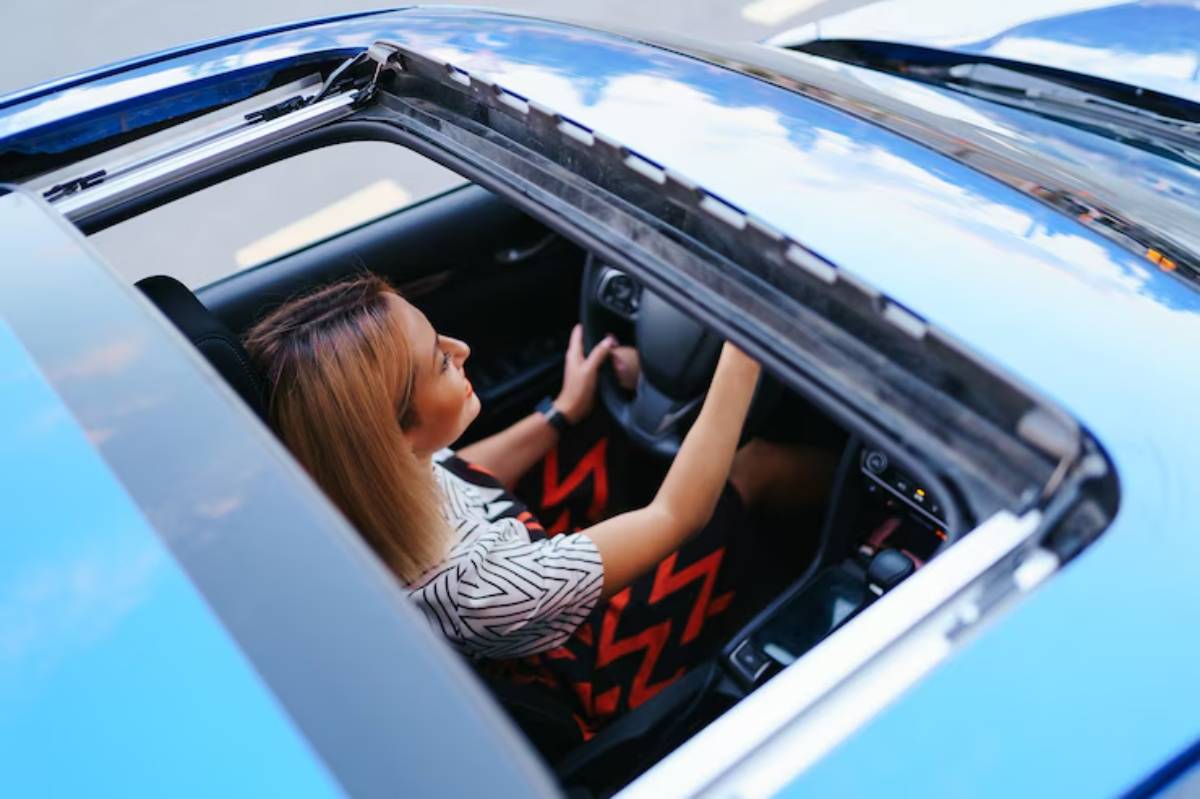 Aerial view of a person driving a blue car with a sunroof, wearing a patterned top and vibrant red skirt.