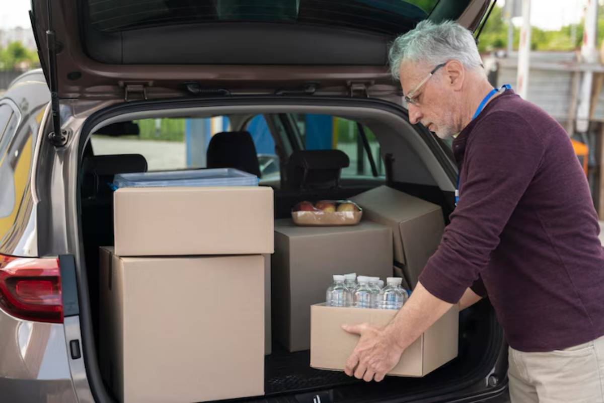 An elderly man loads boxes and bottles of water into the trunk of a grey SUV, surrounded by greenery.