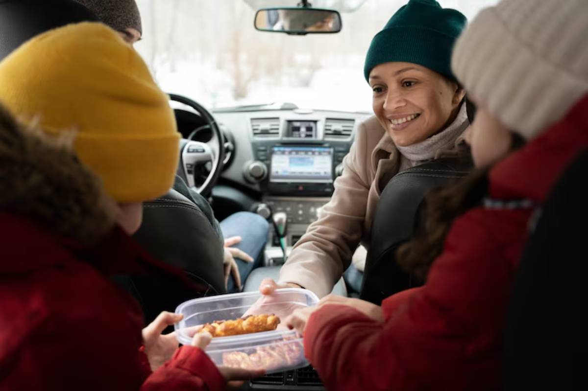 A group of four people in a car, sharing food from a container on a snowy day, with warm clothing and a cozy atmosphere.