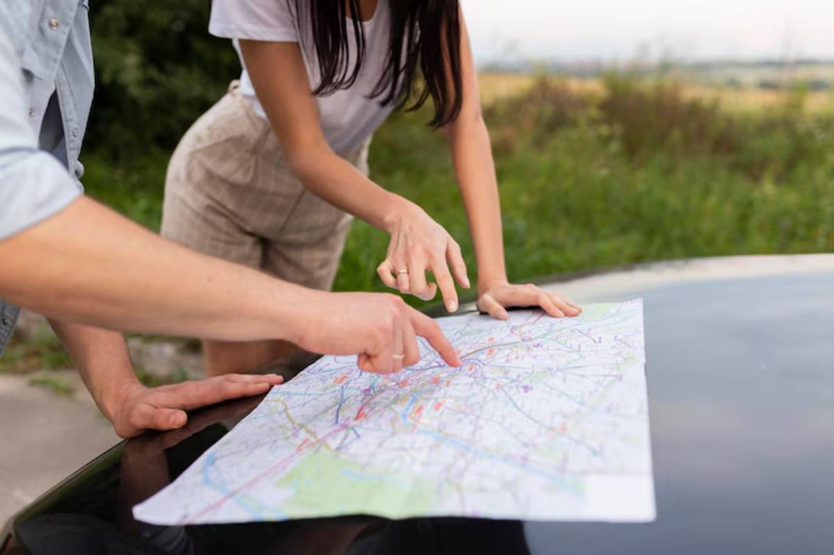 A couple studies a map on a car hood, pointing at locations while surrounded by green fields under an overcast sky.