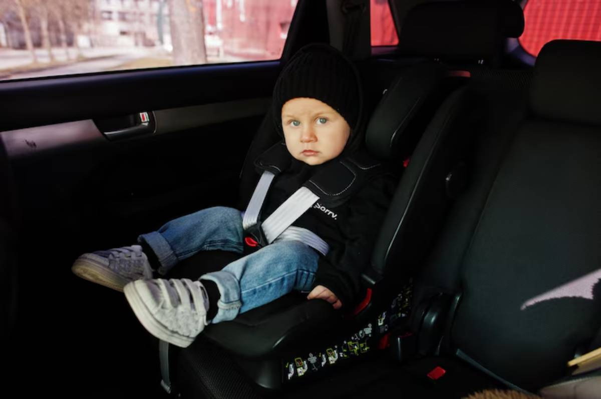 A child in a black car seat wearing a black hoodie and jeans, secured with seatbelts, inside a car.