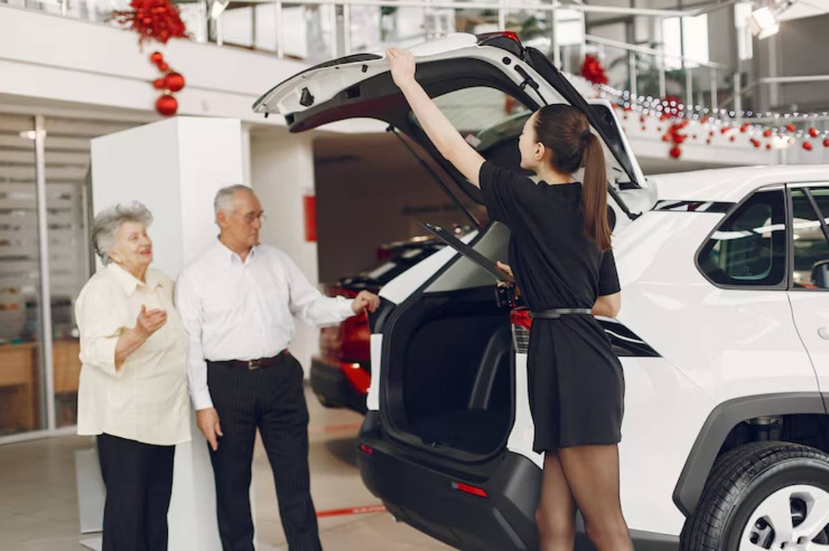 A salesperson in a black dress presents the trunk of a white car to an elderly couple in a festive car showroom.