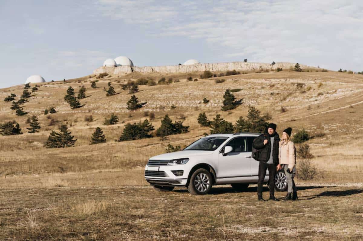 A silver SUV parked on a grassy hillside, with small trees and domes on the distant hill under a partly cloudy sky.