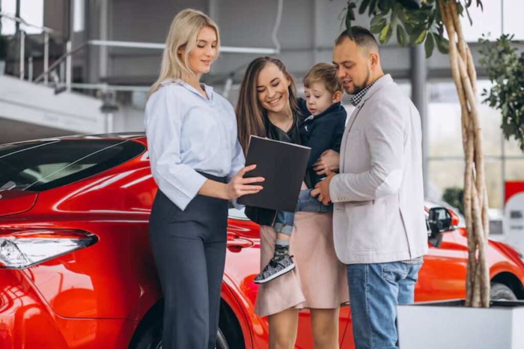 A family interacts with a salesperson at a car dealership, discussing a vehicle next to a sleek red car.