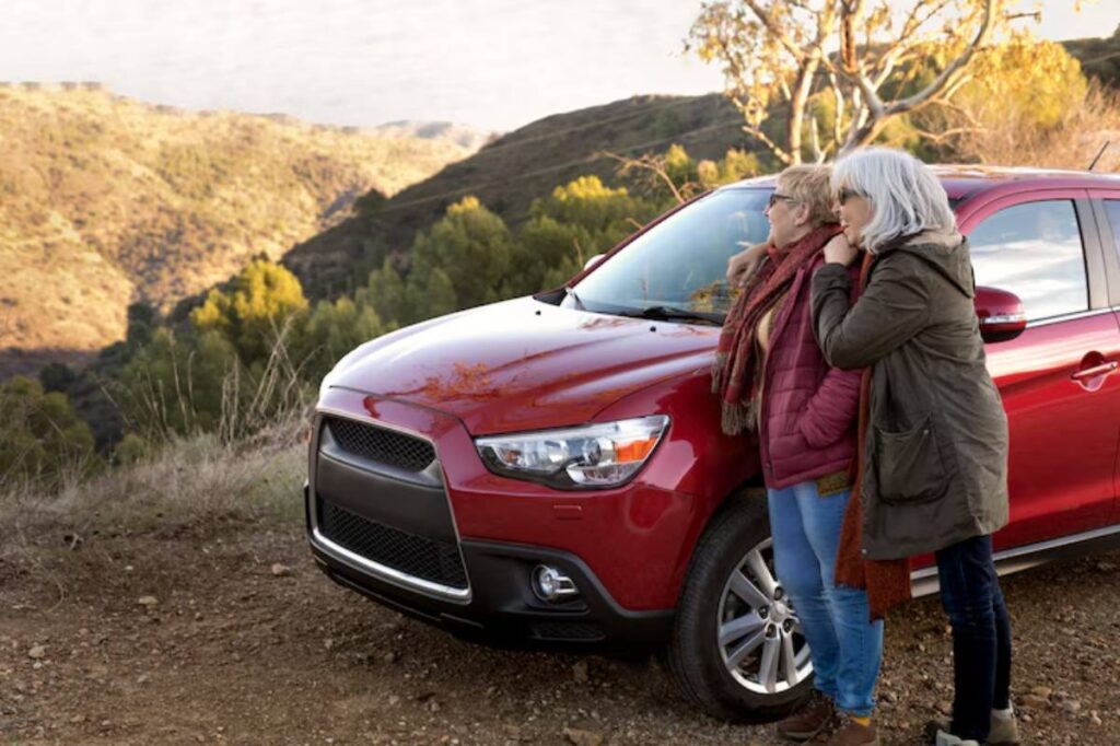 Two women embrace near a red SUV, set against a scenic mountain backdrop with lush greenery and a clear sky.
