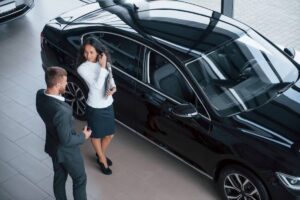 A salesperson in a suit engages with a customer near a sleek black car in a modern showroom.