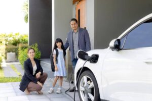 A family stands outside their modern home, next to a white electric vehicle being charged, with lush greenery in the background.