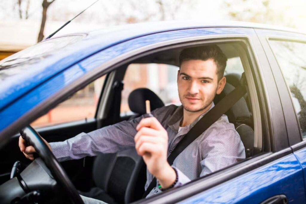 A person sitting inside a blue car holds a car key, wearing a seatbelt, with a blurred background of trees and buildings.