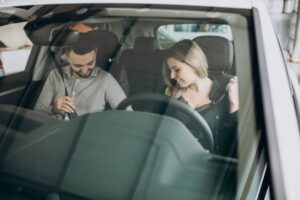 Two individuals sit inside a car, one adjusting their seatbelt while the other leans towards the steering wheel. Natural light filters in.