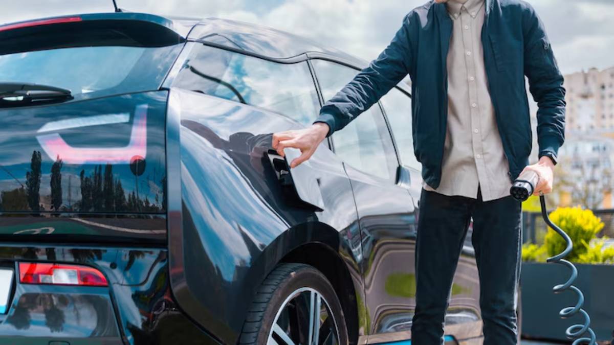 A person in a jacket plugs an electric car into a charging station, surrounded by greenery and a clear sky.