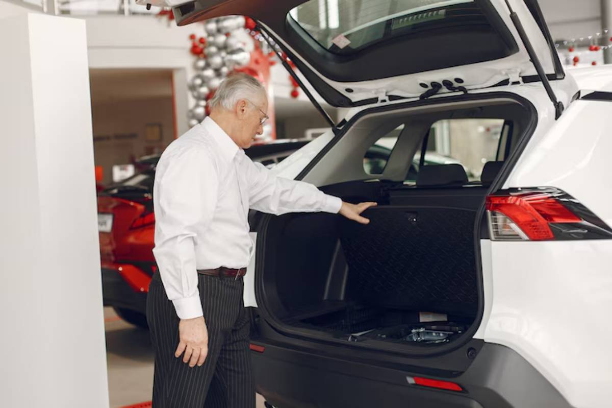 An elderly man in a white shirt examines the trunk of a white SUV in a car dealership with festive decorations in the background.