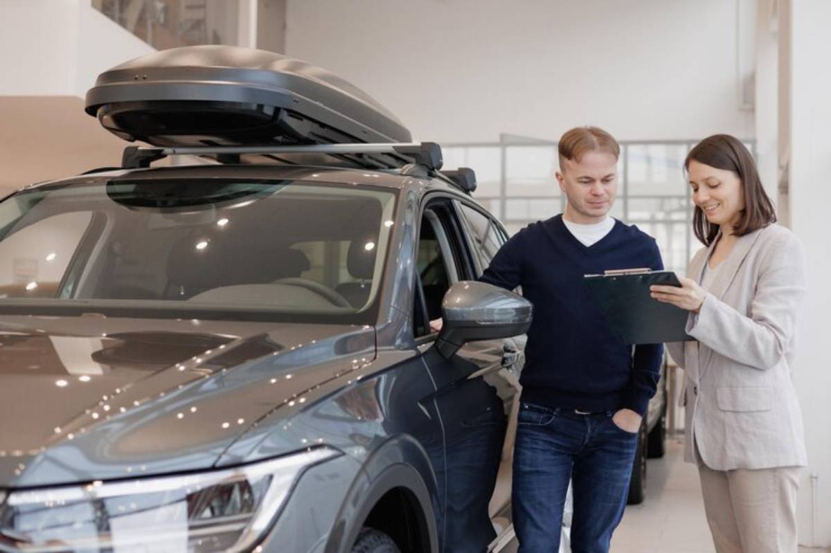 A man and woman discuss a car's features inside a bright, modern dealership, with a roof box mounted on the vehicle.