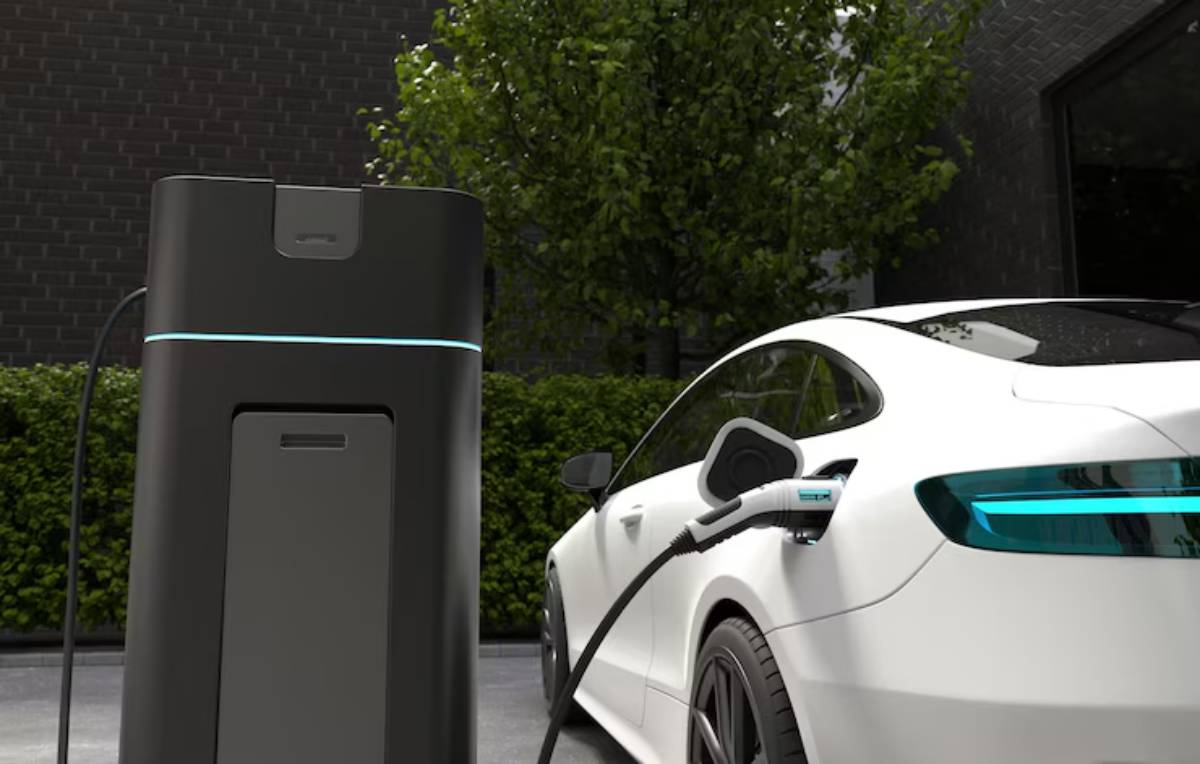 A sleek white electric car is charging at a modern black electric vehicle charger, set against lush greenery and a dark brick background.