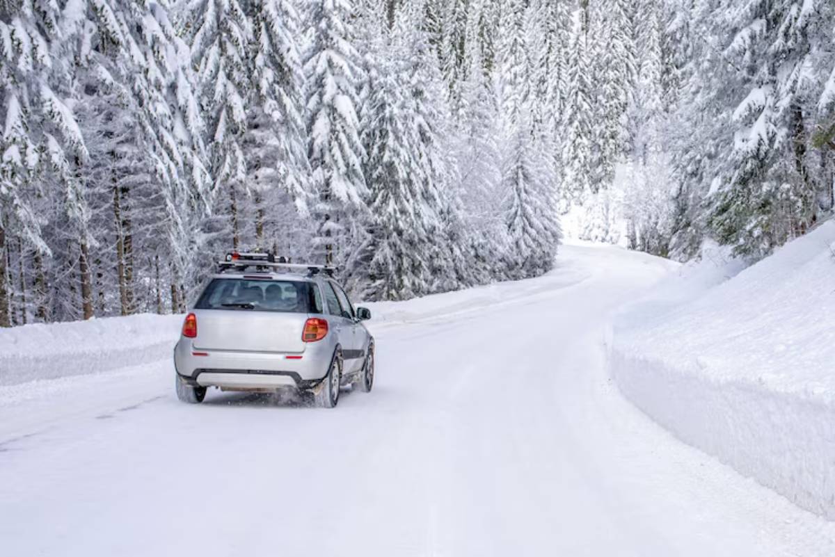 A silver car drives on a snowy road surrounded by tall, snow-covered trees in a serene winter landscape.