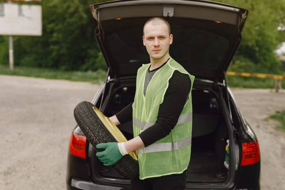A person in a reflective safety vest holds a tire while standing next to an open car trunk in a green outdoor setting.