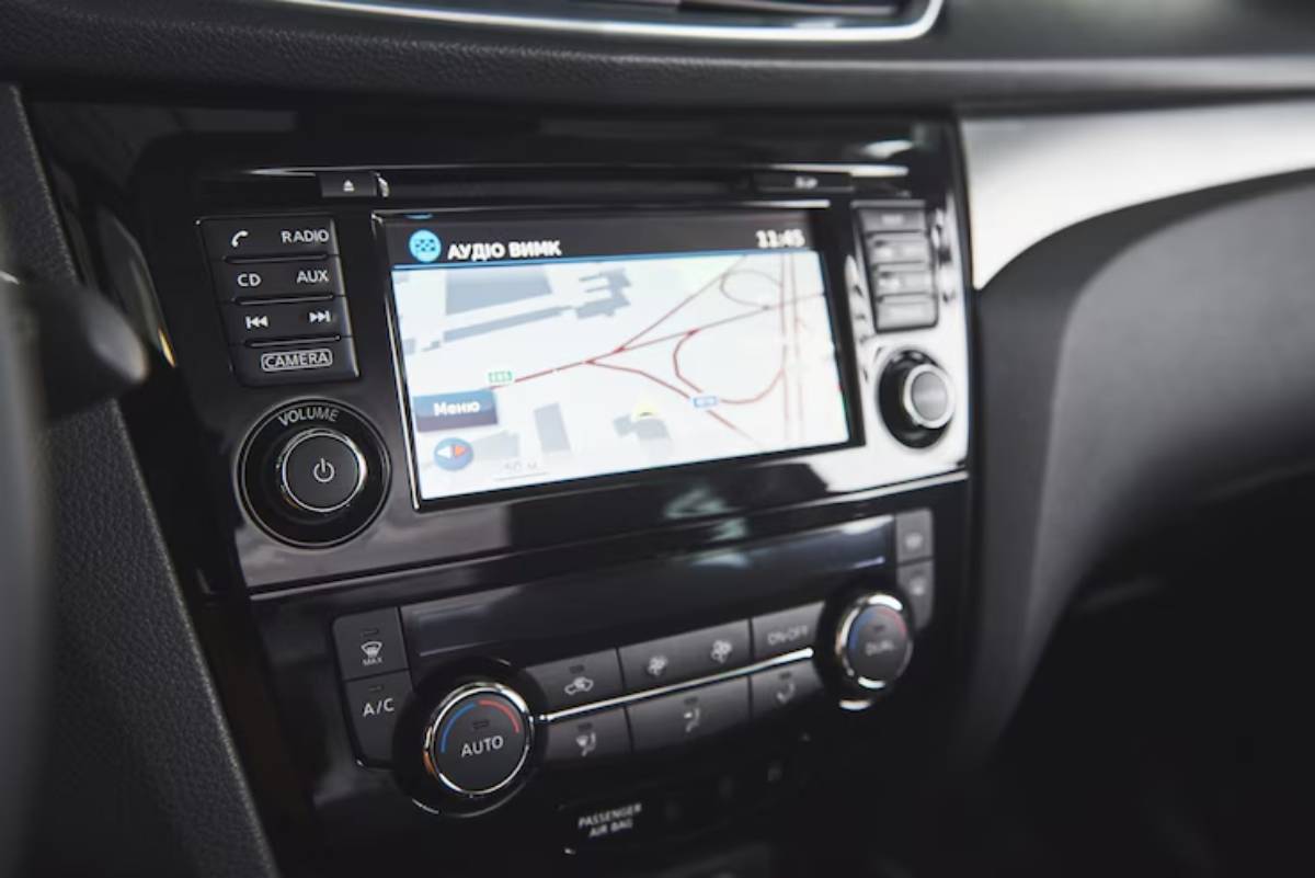 A driver's hand on the wheel with a GPS screen displaying navigation directions on a sunny road surrounded by autumn trees.