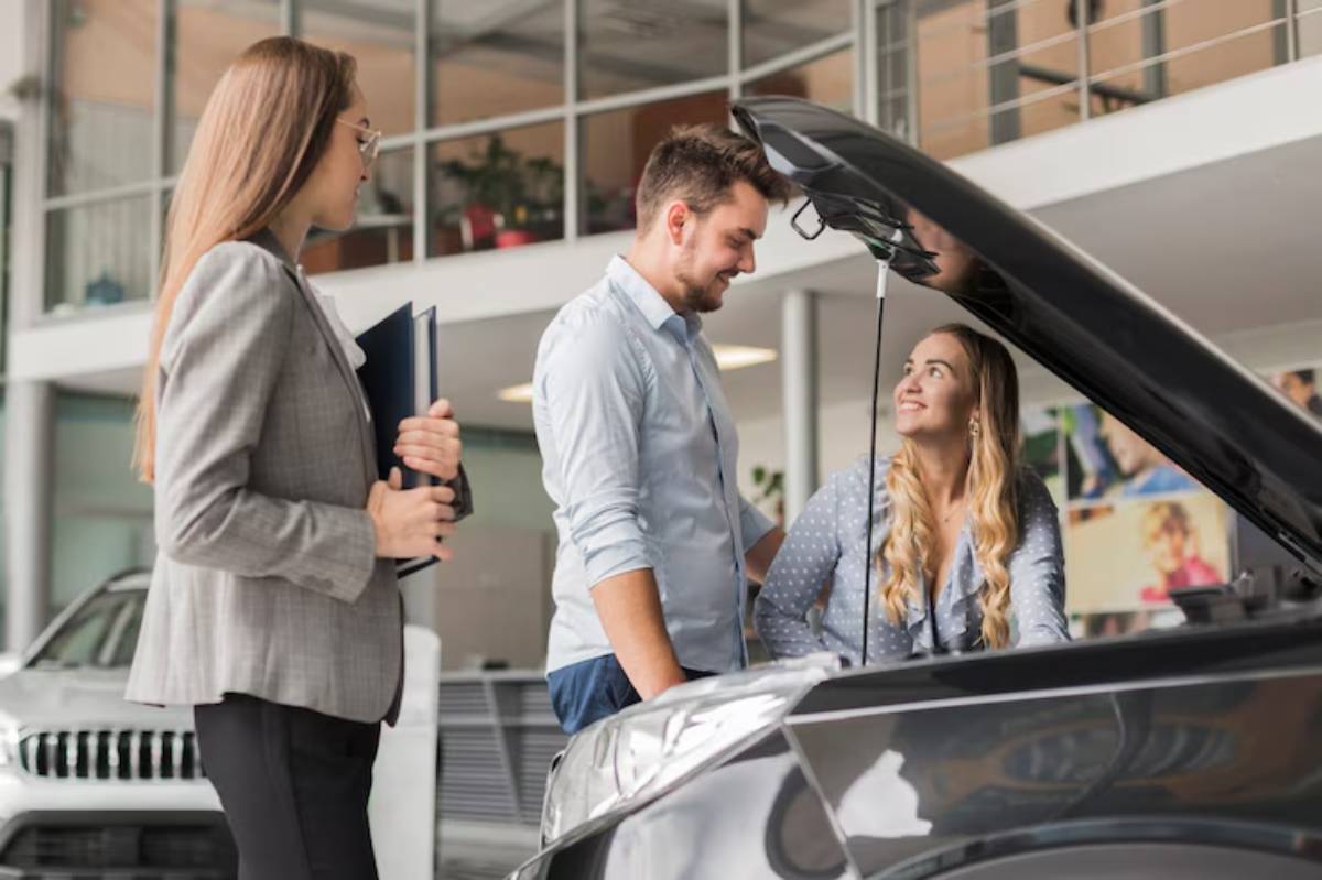 A woman in a blazer shows a notebook to a man examining a car engine with another woman beside him in a dealership setting.