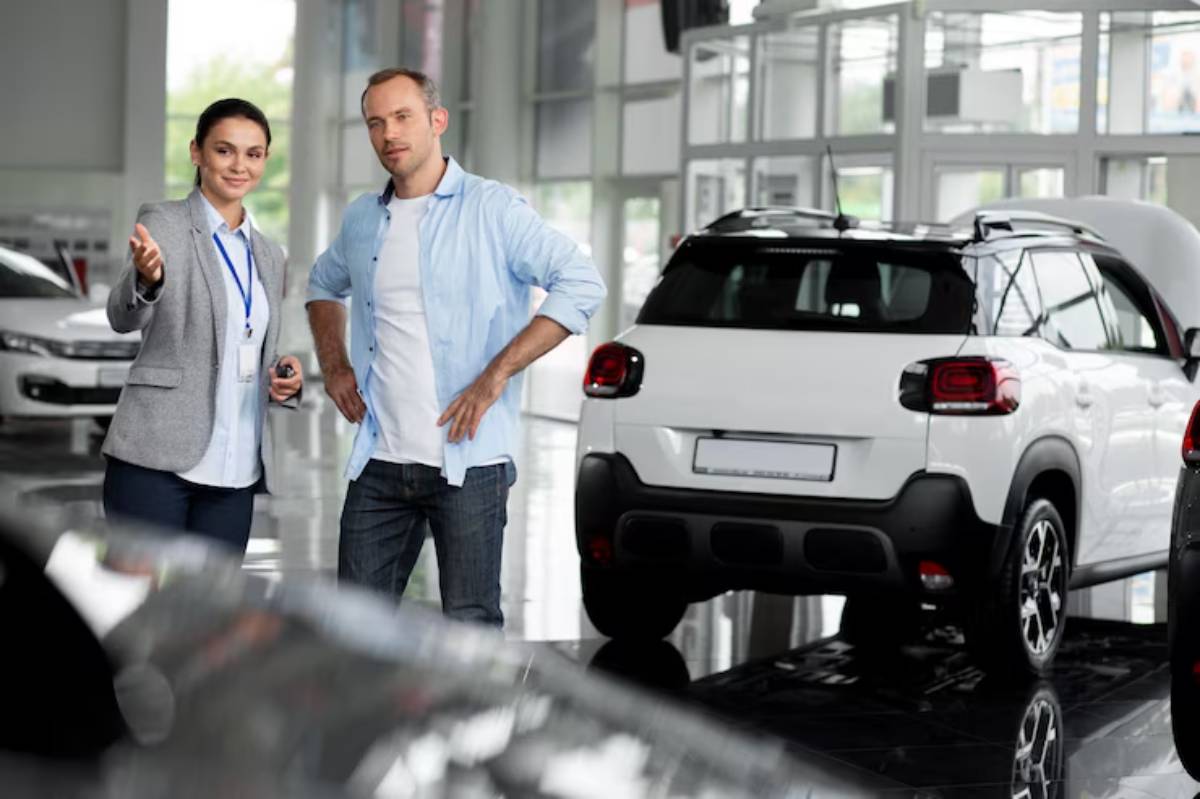 A car dealership scene with a salesperson gesturing while discussing a vehicle with a customer, featuring a white car in the background.