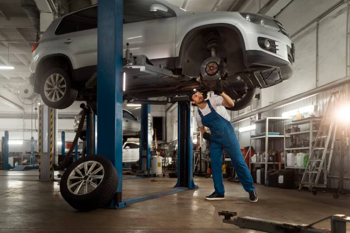 A mechanic in blue overalls works under a lifted silver SUV in a well-equipped automotive workshop, surrounded by tools and equipment.